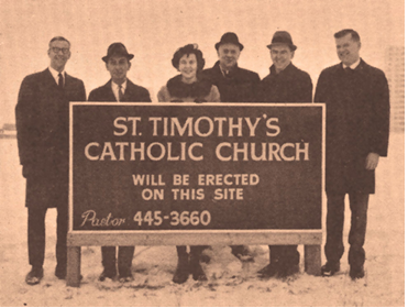 Six People holding a sign saying "St Timothy's Catholic Church will be erected in this site."  The sign included the Pastors telephone number.